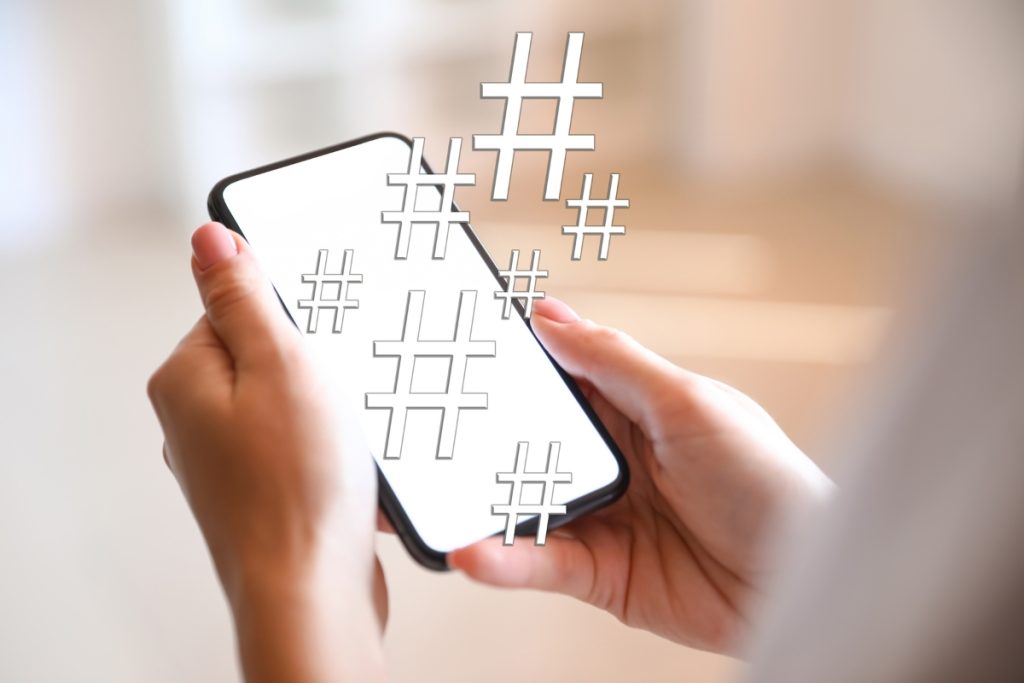 A close-up of a person's hands holding a smartphone with several translucent, digital hashtag symbols floating off the screen.
