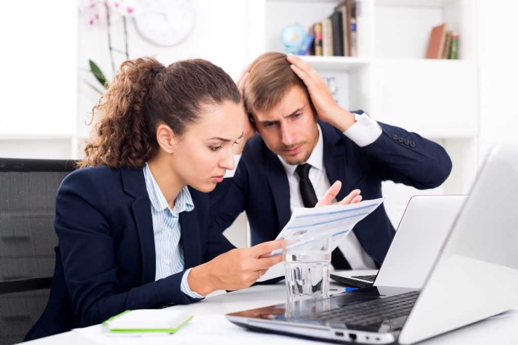 A stressed businesswoman and businessman at a desk looking at a document. The man has his hands on his head in frustration while looking over her shoulder at the paperwork near a laptop.