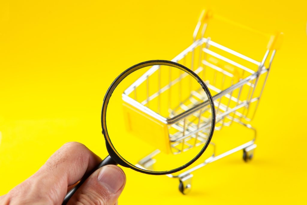A hand holding a magnifying glass over a small, empty silver and yellow toy shopping cart against a bright yellow background.