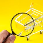 A hand holding a magnifying glass over a small, empty silver and yellow toy shopping cart against a bright yellow background.