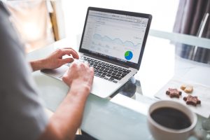 An over-the-shoulder view of a person typing on a laptop at a glass table. The laptop screen displays a web analytics dashboard featuring data line graphs and a pie chart next to a cup of coffee and cookies.