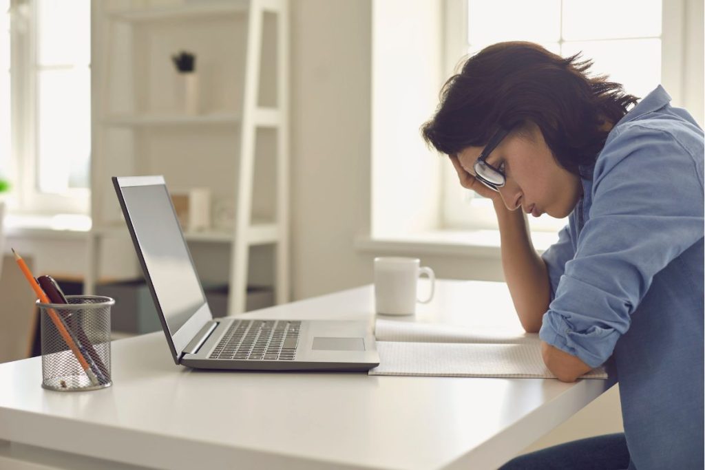 A frustrated woman sitting at a desk resting her head in her hand while staring down next to an open laptop.
