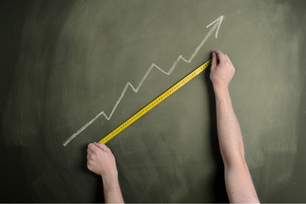A pair of hands using a yellow measuring tape along an upward-trending chalk arrow drawn on a green chalkboard.