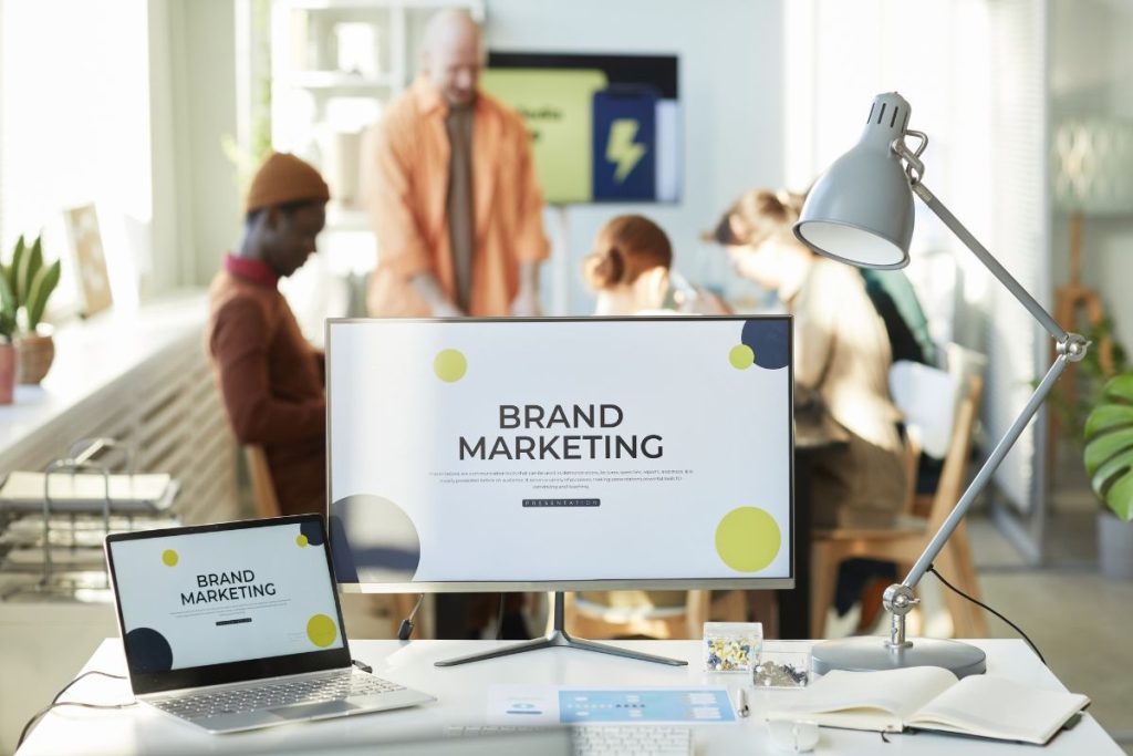 A modern office workspace with a computer monitor in the foreground showing a "BRAND MARKETING" presentation slide.