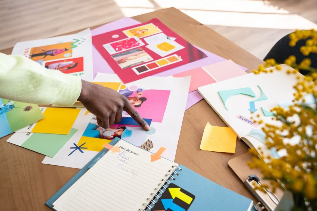 An overhead view of a designer's desk with hands pointing to colourful mood boards, swatches, and a notebook.